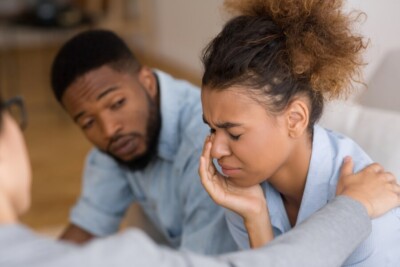 Photo of a woman crying, a man looking at her and another woman putting a hand on her shoulder. The couples is black. This photo represents the betrayal trauma that can happen after an affair. If you're looking for an affair counselor in Georgia, we can help.
